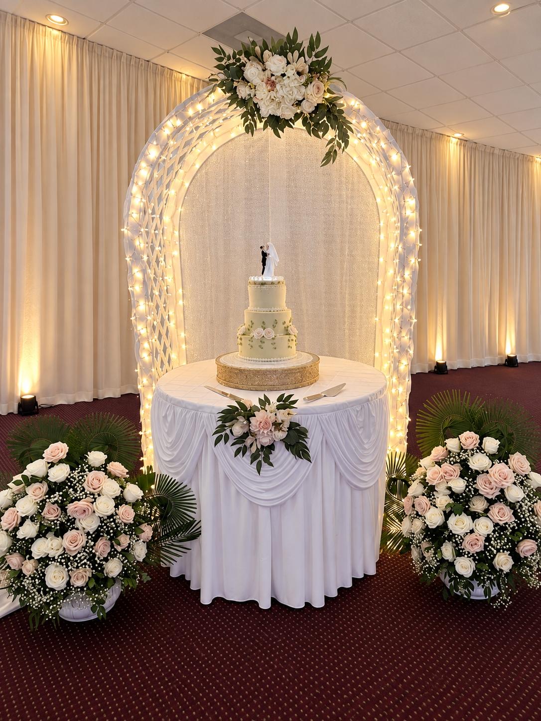 Wedding cake displayed under an illuminated white arch with flowers